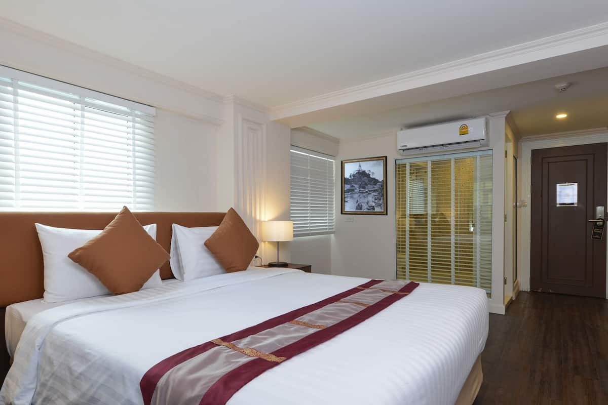 A hotel room with a large bed, a headboard, a nightstand, a lamp, a window with blinds, a door, and an air conditioning unit.