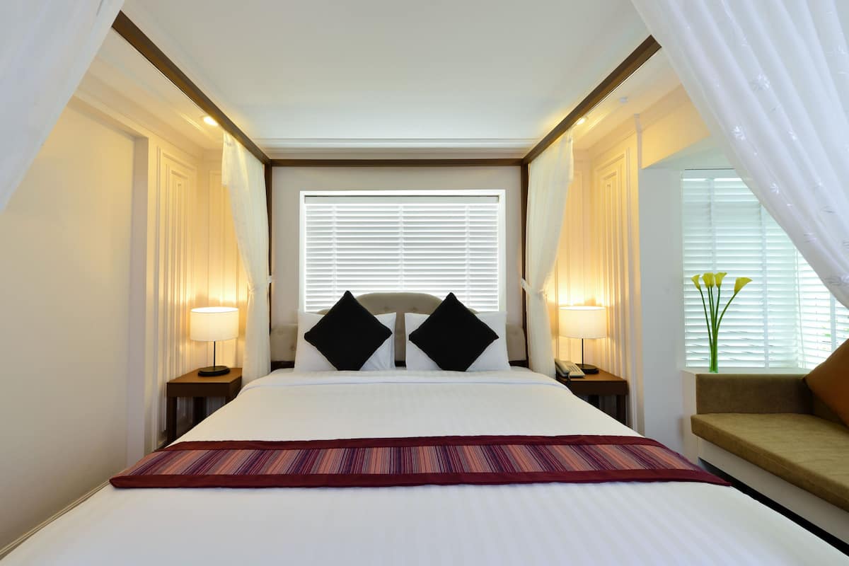 A hotel room with a large bed, two bedside tables with lamps, a window with blinds, and a small seating area with a cushion.