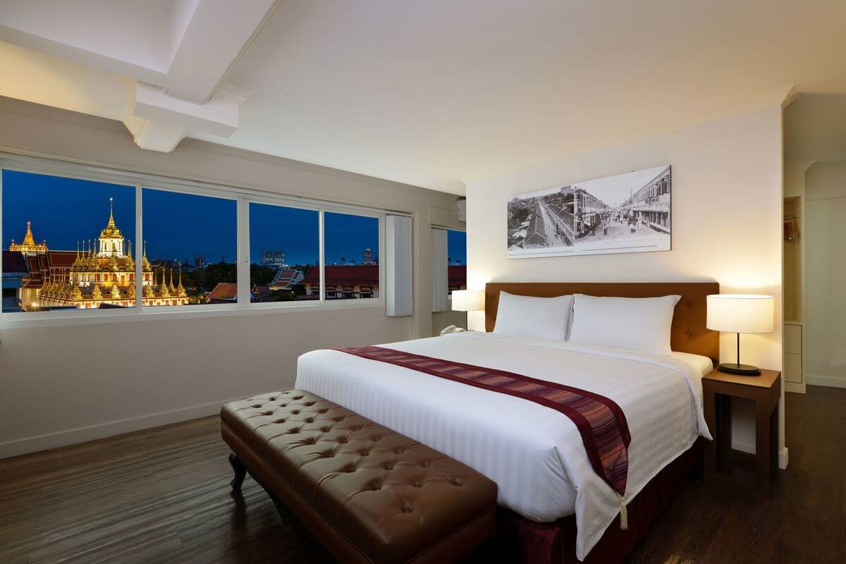 A hotel room with a large bed, a bench, a nightstand, a lamp, and a window with a cityscape view.
