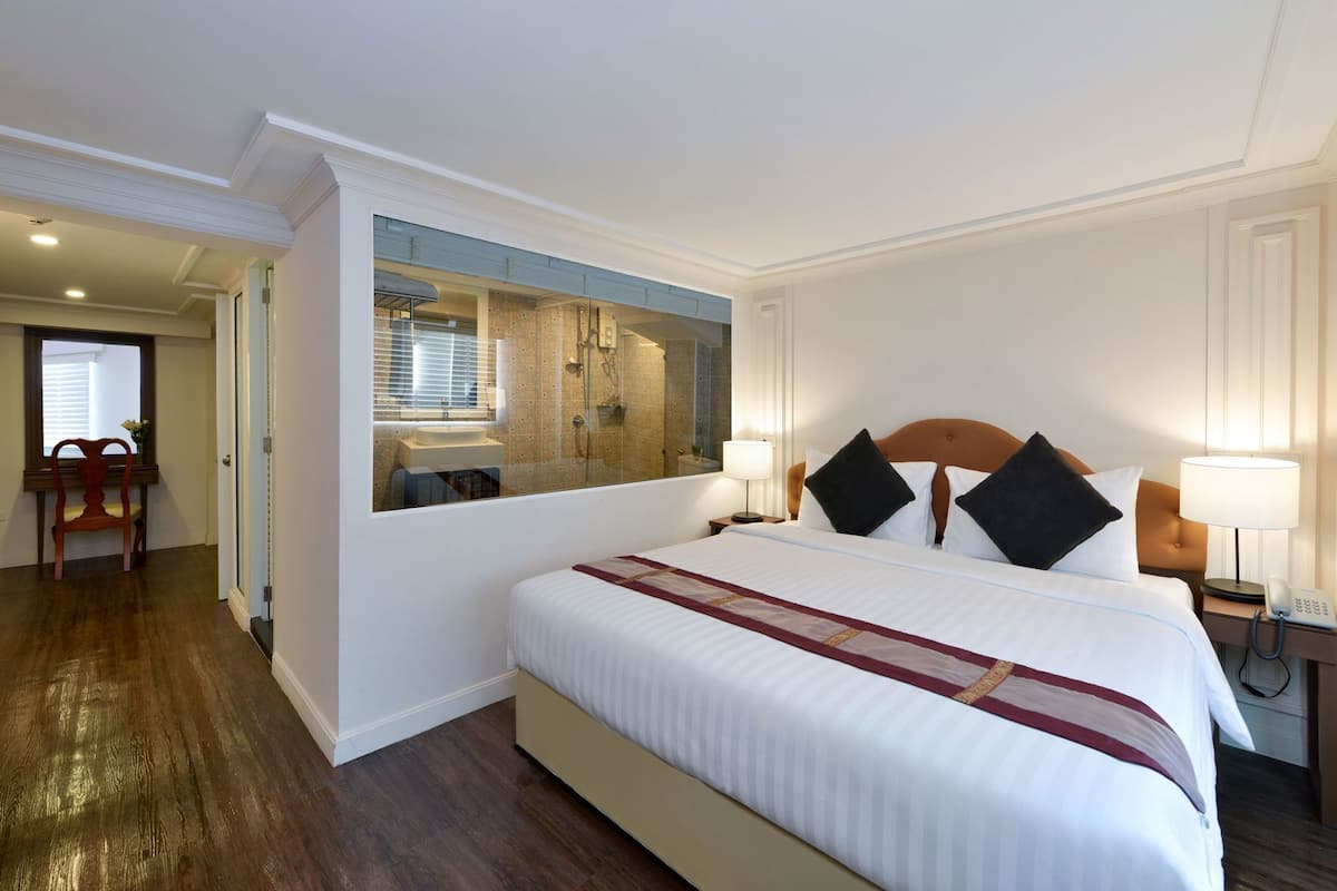 A hotel room with a large bed, a mirror, a bathroom visible through the mirror, a desk with a lamp, and a chair.