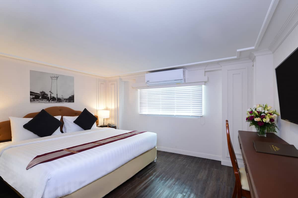 A hotel room with a bed, bedside table, lamp, air conditioning unit, window with blinds, and a flat-screen TV.