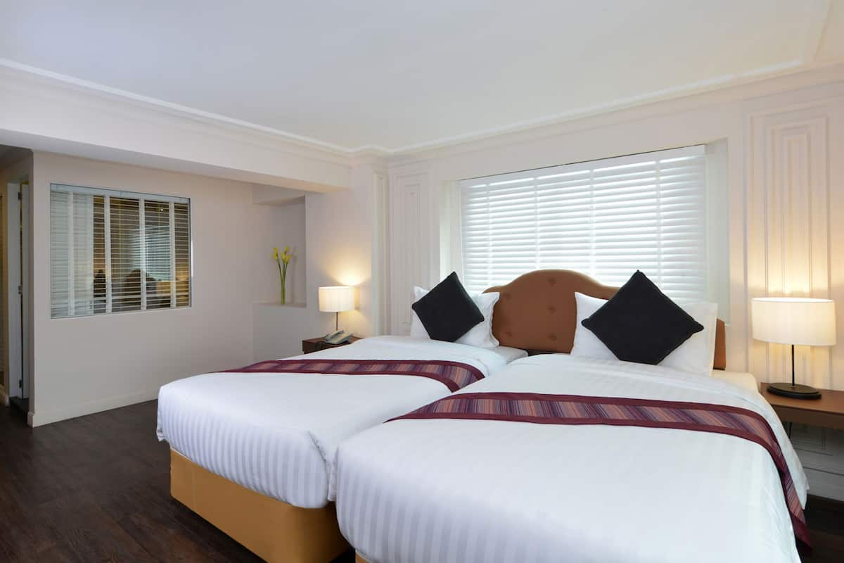 A hotel room with two beds, a nightstand with a lamp, a window with blinds, and a door leading to another room.
