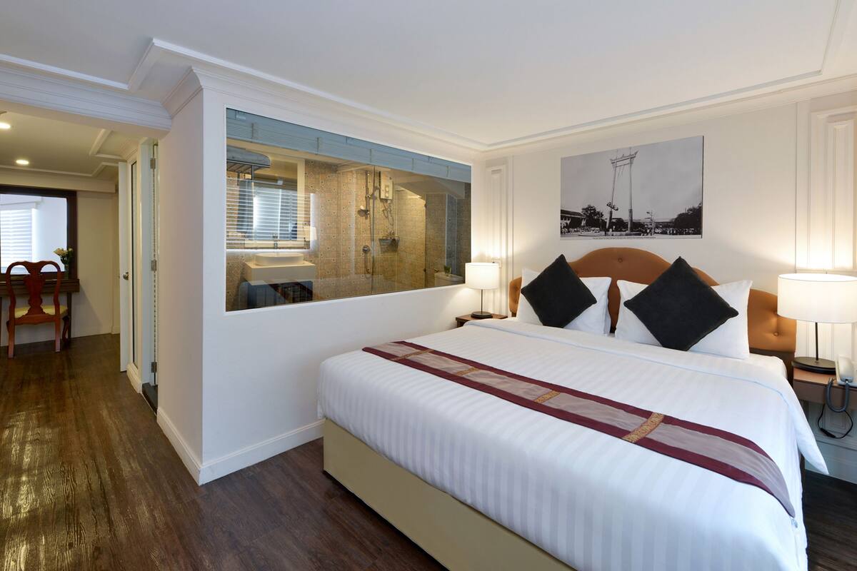 A hotel room with a large bed, a bathroom with a glass shower, a wooden chair, and a painting on the wall.