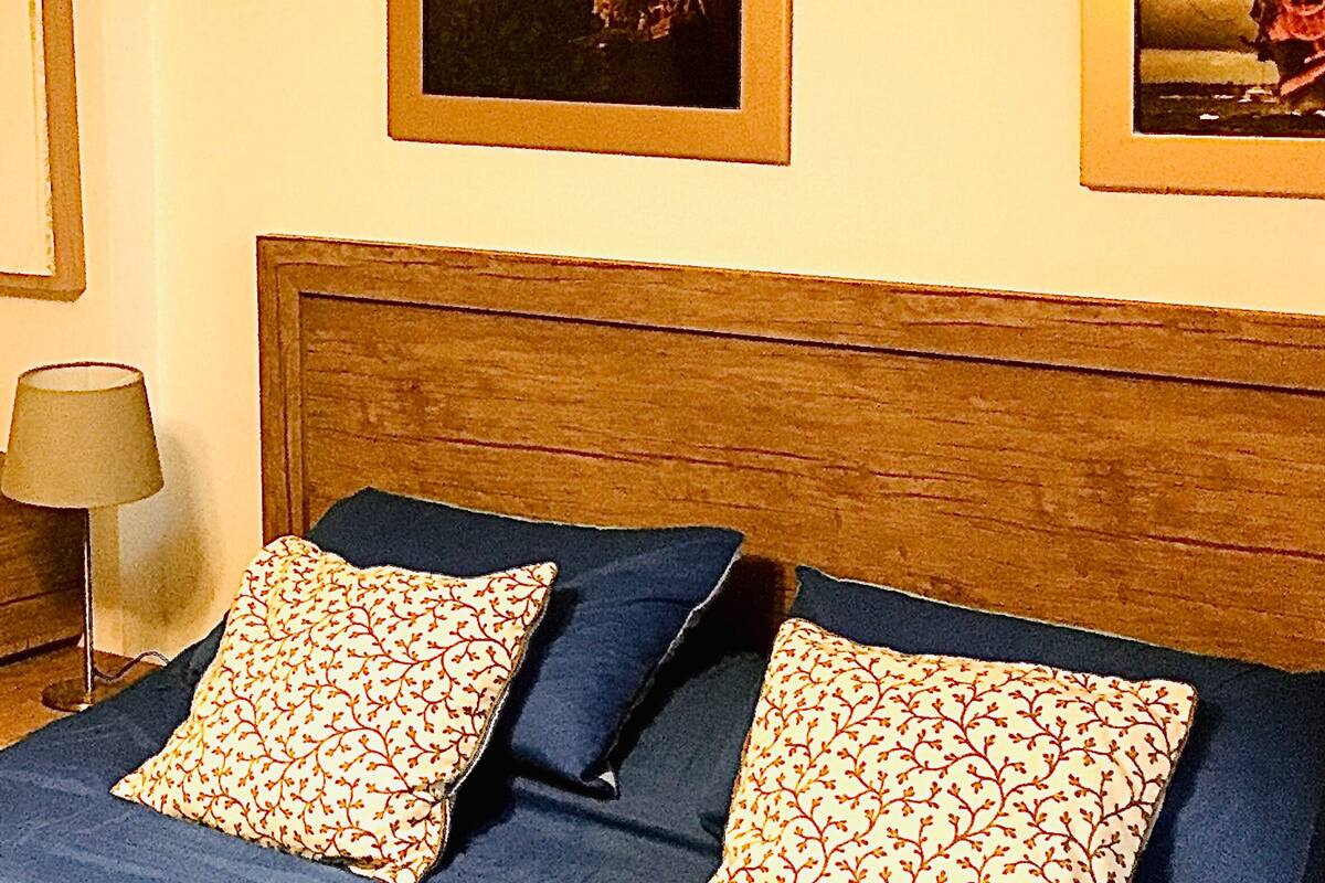 A bed with blue bedding and patterned pillows, a wooden nightstand with a lamp, and a small wooden box.