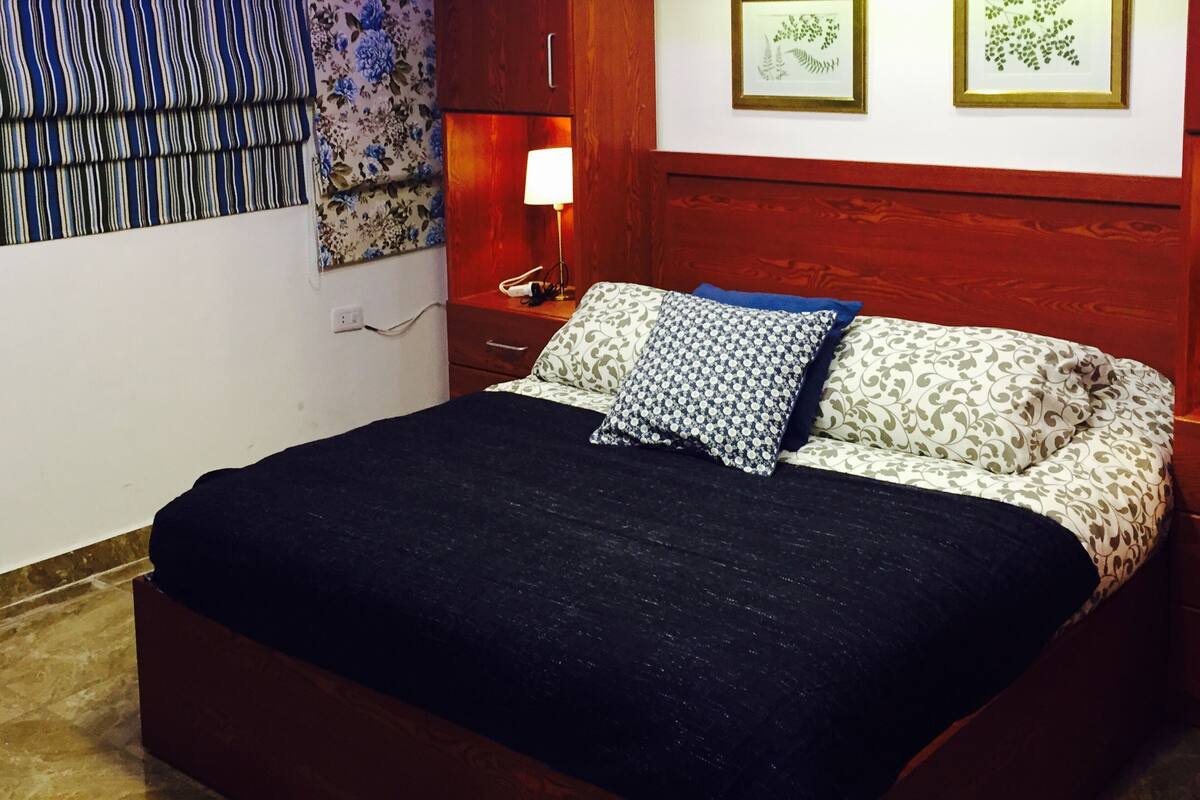 A bedroom with a bed, a nightstand, a lamp, and two framed botanical prints on the wall.