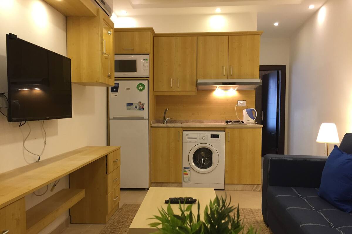 A compact kitchen with wooden cabinets, a microwave, a refrigerator, and a washing machine. There is a sofa and a TV mounted on the wall.