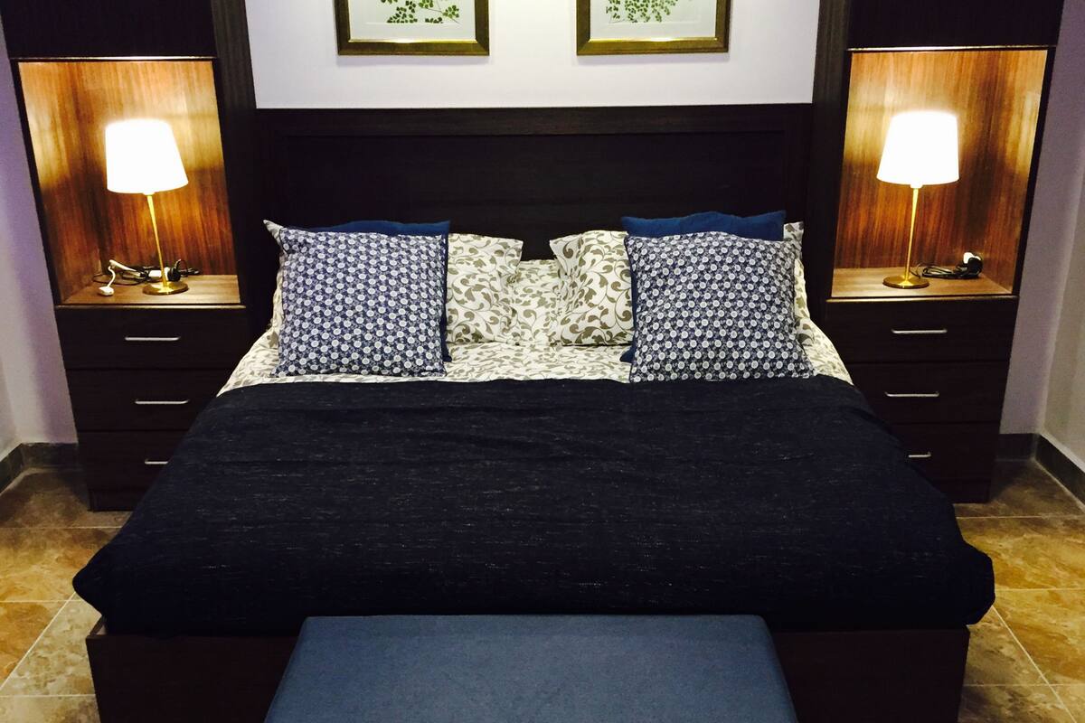 A neatly made bed with a black bedspread and matching bench, flanked by patterned pillows and two bedside tables with lamps.
