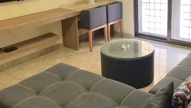 A modern living room with a flat-screen TV, a glass coffee table, a sofa, and a small dining area with a plant.