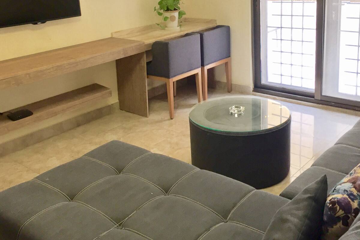 A modern living room with a flat-screen TV, a glass coffee table, a sofa, and a small dining area with a plant.