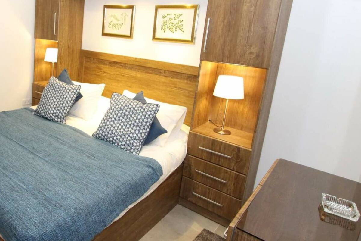 A hotel room with a large bed, wooden headboard, two bedside tables with lamps, a small wooden nightstand, and a mirror.