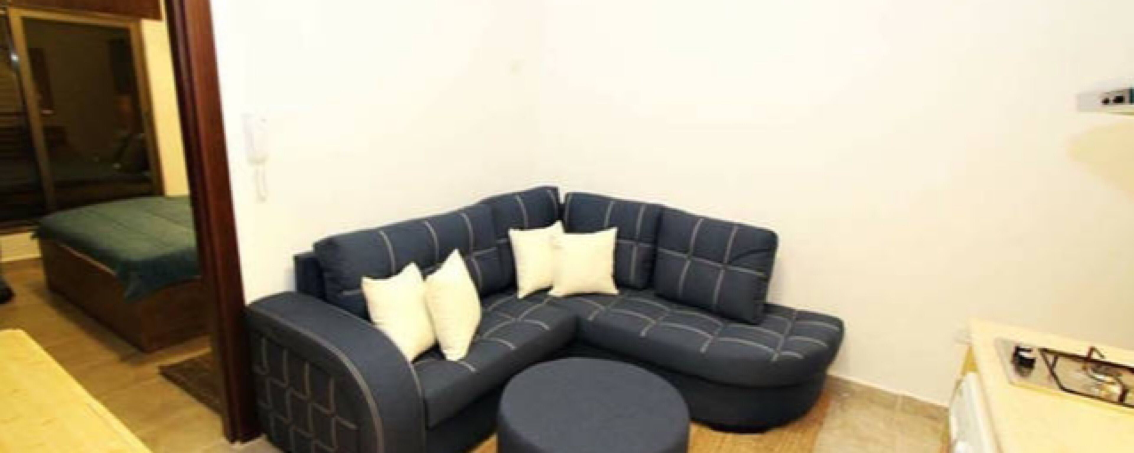 Una sala de estar moderna con un sofá seccional oscuro, un otoman redondo y una mesita con una lámpara.