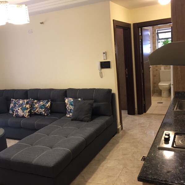 A modern living room with a dark grey sectional sofa, a small round table, and a built-in kitchen area with a black countertop and stove. - The Royal Luxury Apartments & studios (Amman)