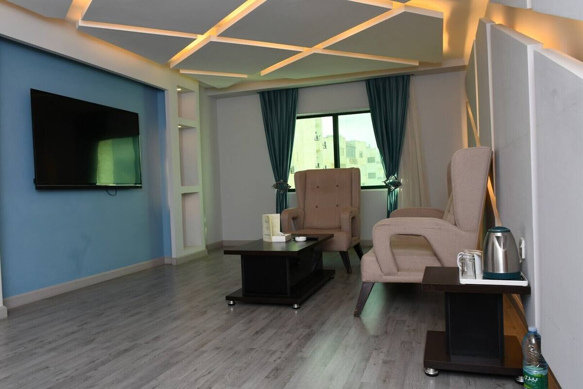 A modern living room with a flat-screen TV, two armchairs, a coffee table, and a side table with a kettle and water bottle.