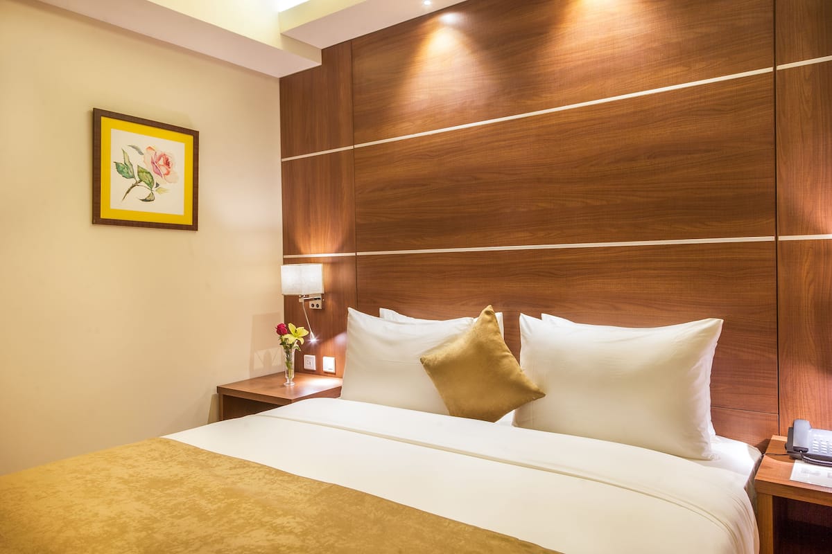 A hotel room with a wooden headboard, a bed with white linens, a gold throw blanket, a bedside table with a lamp and a vase of flowers, and a framed picture on the wall.