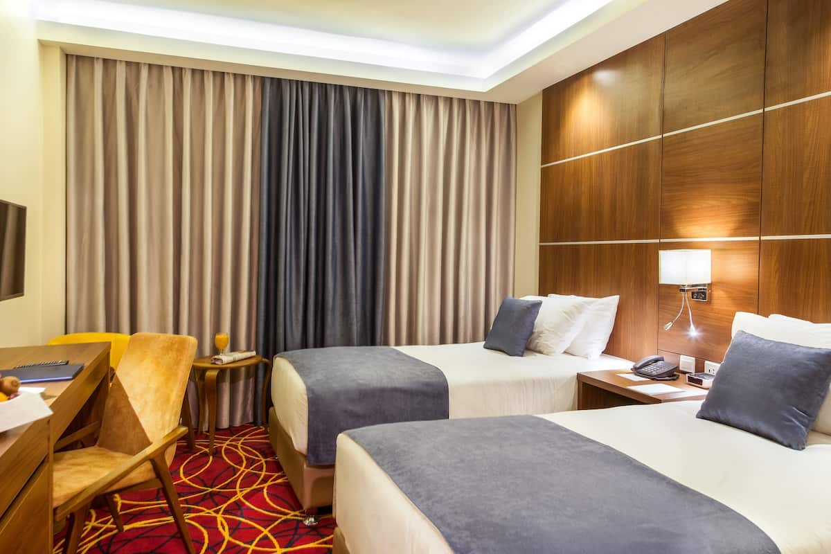 A hotel room with two beds, a desk, a chair, a small table with a glass of orange juice, and a lamp on the nightstand.