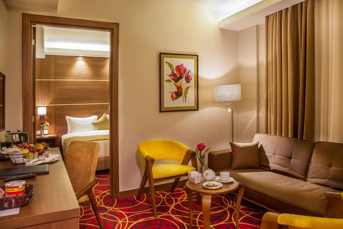 A hotel room with a bed, a yellow armchair, a sofa, a small table with a vase of flowers, a lamp, and a painting of flowers on the wall.