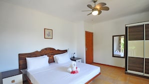 A neatly made bed with white linens and a towel arrangement, a wooden headboard, a nightstand with a lamp, a ceiling fan, and a wardrobe with a mirror. - GT Self Catering Apartments (Mahé Island)