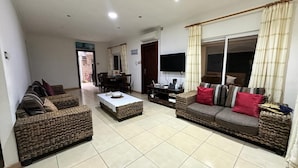 A living room with wicker sofas, a coffee table, a flat-screen TV, and a dining area with chairs and a table. - GT Self Catering Apartments (Mahé Island)