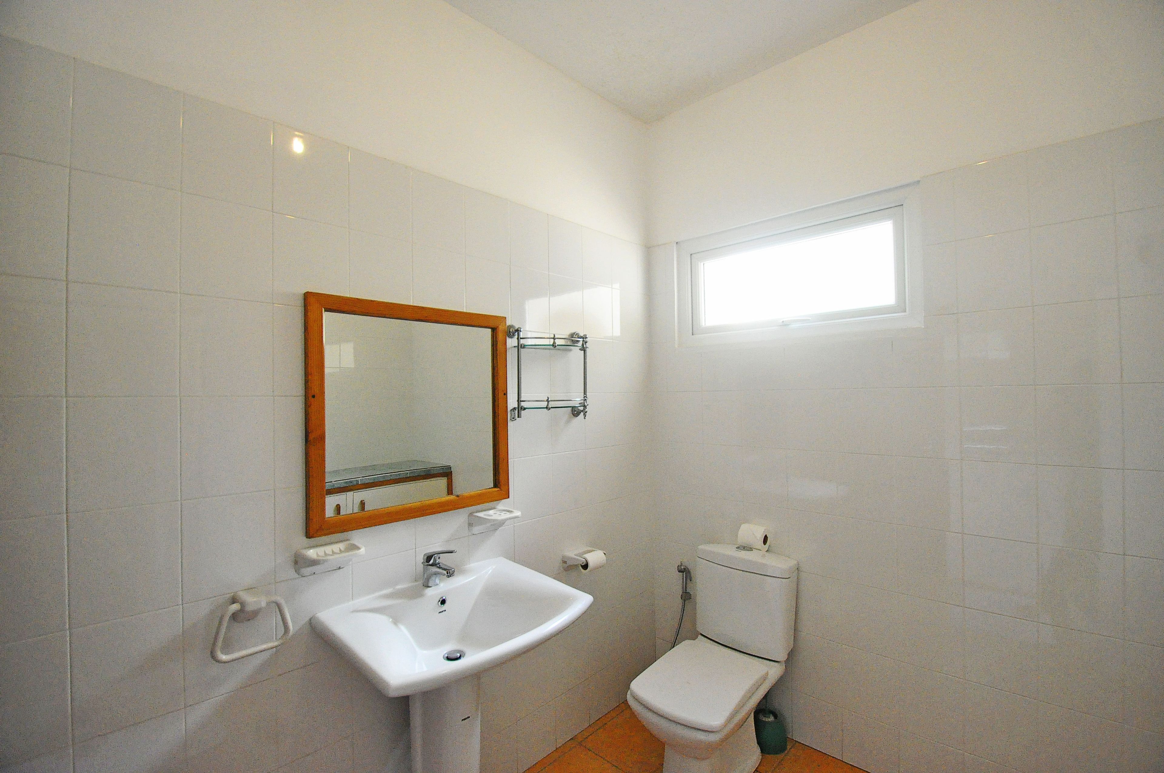 A small, clean bathroom with white tiles, a sink, a toilet, and a window providing natural light.