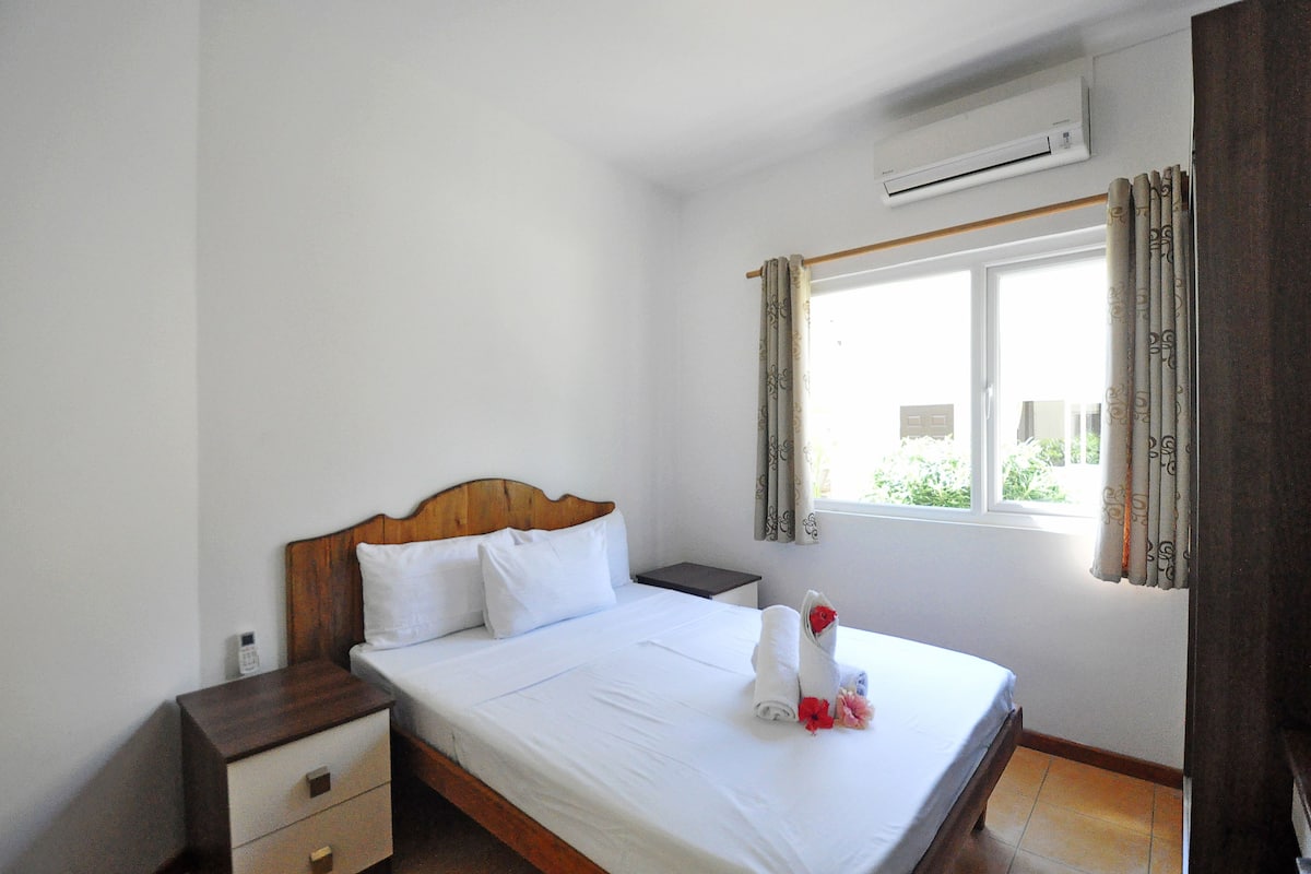 Habitación de hotel con cama de madera, ropa de cama blanca, una mesita de noche, un cajón pequeño y una ventana con cortinas.
