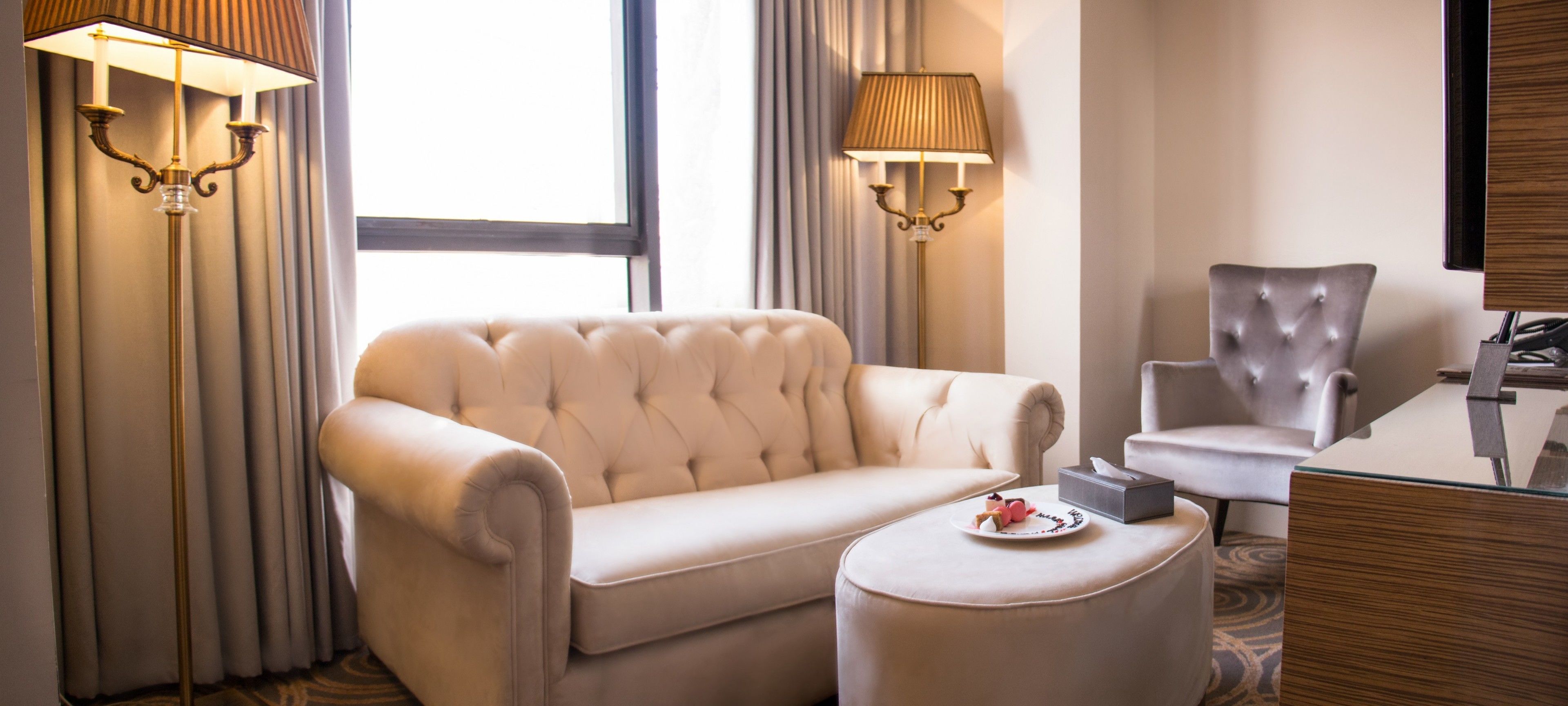 A hotel room with a tufted sofa, a matching ottoman, a dining table with a plate of fruit, and two wall-mounted lamps.