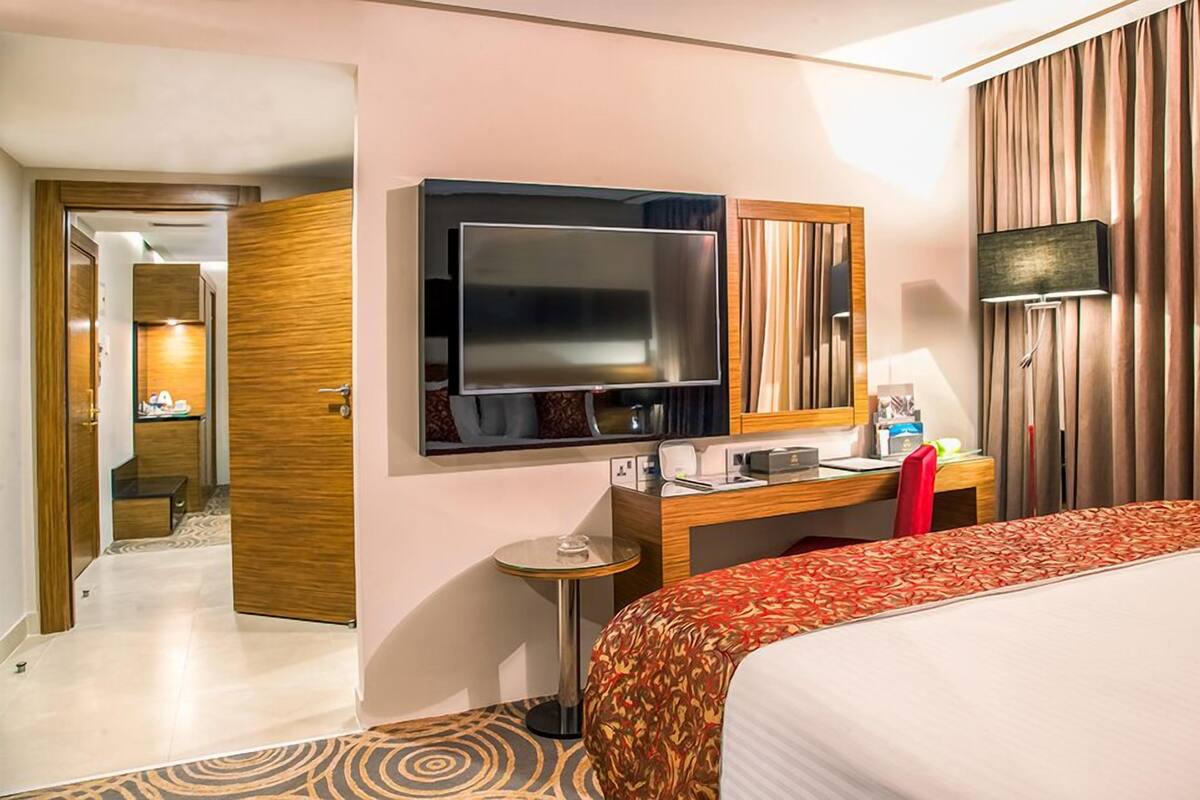 A hotel room with a bed, a desk with a computer, a TV, a mirror, a lamp, and a bathroom visible through an open door.