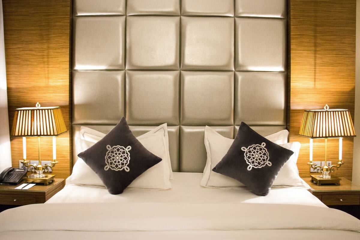 A hotel room with a large padded headboard, two bedside tables with lamps, and decorative pillows.