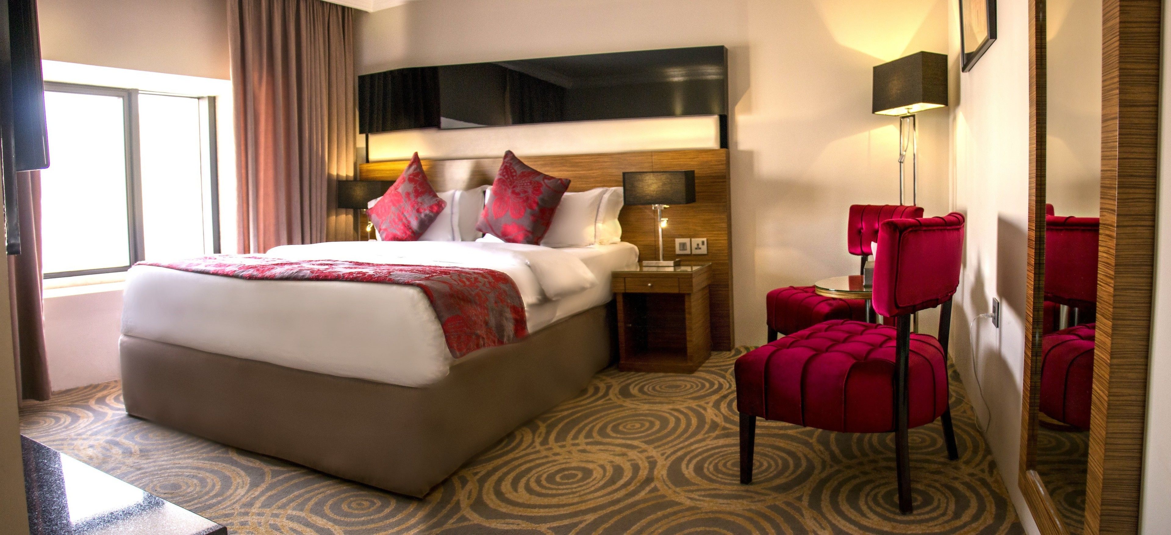 Une chambre d’hôtel avec un grand lit, une table de chevet, une lampe et un coin salon avec des fauteuils rouges.