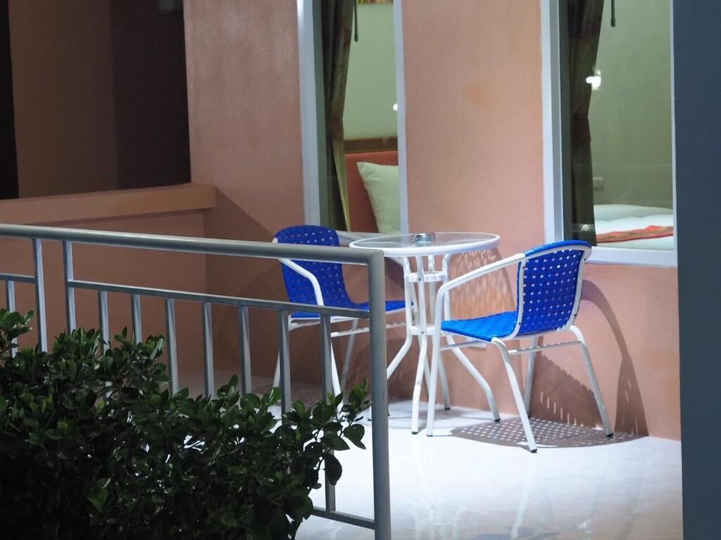 A balcony with two blue chairs and a small table, a metal railing, and a view of a building through a window.