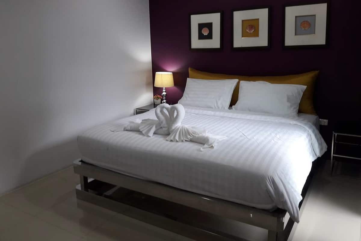 Een netjes opgemaakt bed met witte lakens en een decoratieve handdoekopstelling. Er staat een nachtkastje met een lamp bij en aan de muur hangt een lijst met een afbeelding.