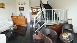 A modern living room with a white sofa, a wooden reception desk, wicker chairs, and a staircase with a glass railing. - David Residence (Mai Khao)