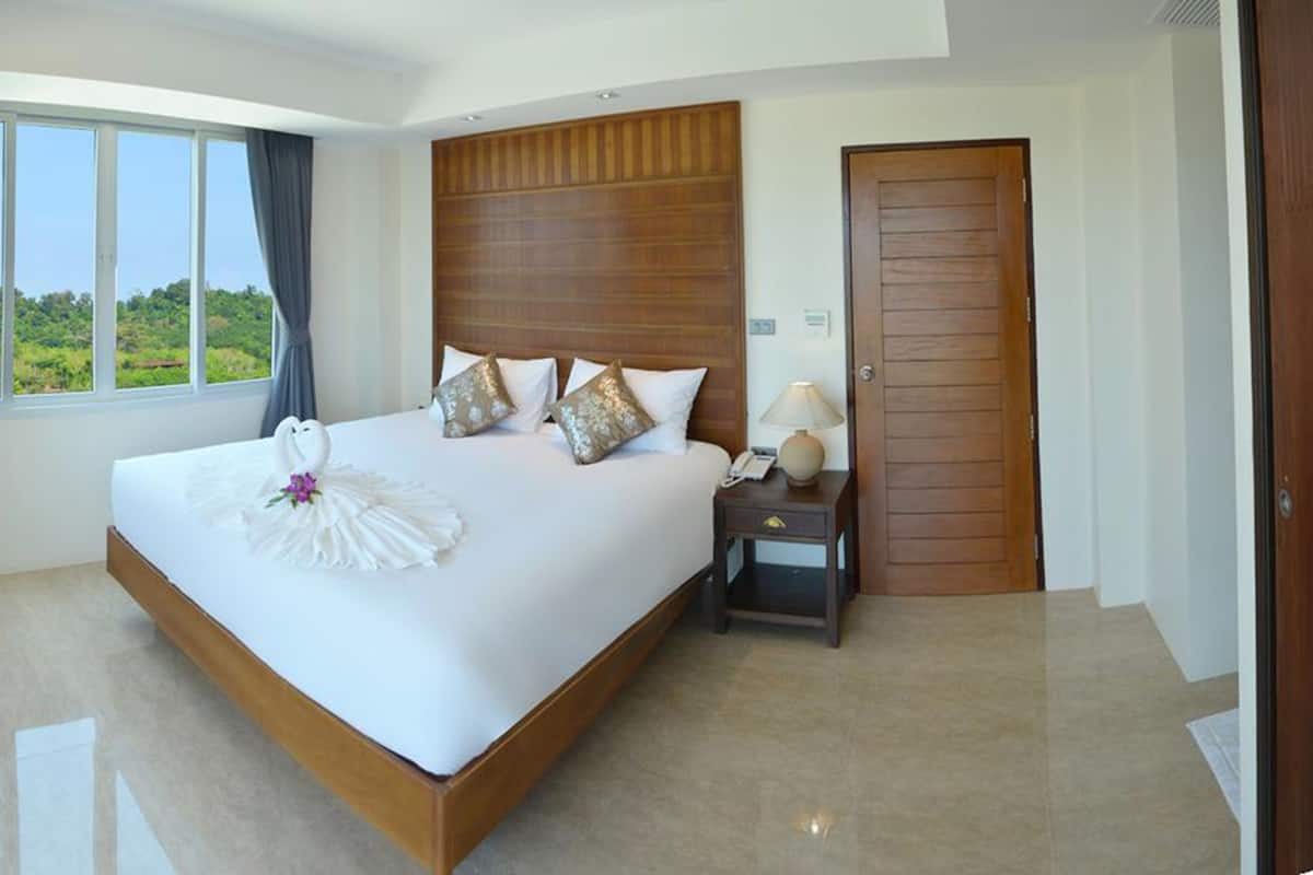 A large bed with a wooden headboard, a bedside table with a lamp, and a view of greenery through the window.