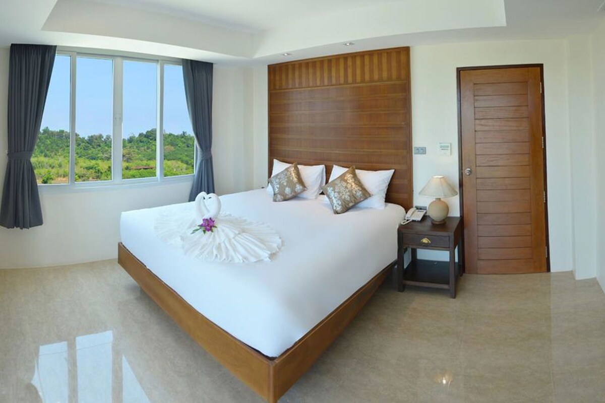 A hotel room with a large bed, a wooden headboard, a nightstand with a lamp, and a view of greenery outside.
