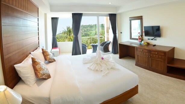 A hotel room with a large bed, a desk, a TV, and a balcony with a view of greenery. - David Residence (Mai Khao)