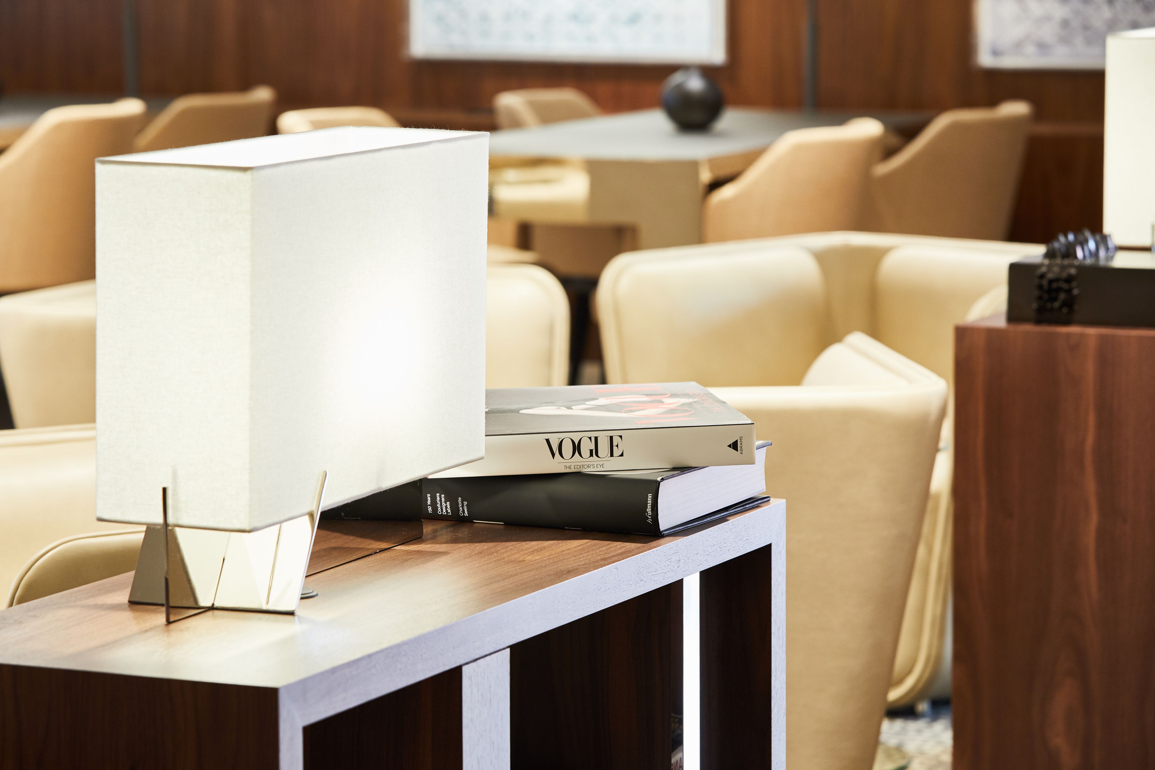 A modern hotel lobby with a wooden reception desk, a white lamp, and a stack of books including a Vogue magazine.