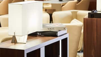 A modern hotel lobby with a wooden reception desk, a white lamp, and a stack of books including a Vogue magazine.