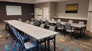 A conference room with long tables and chairs, a projector screen, a storage area, and a carpeted floor.