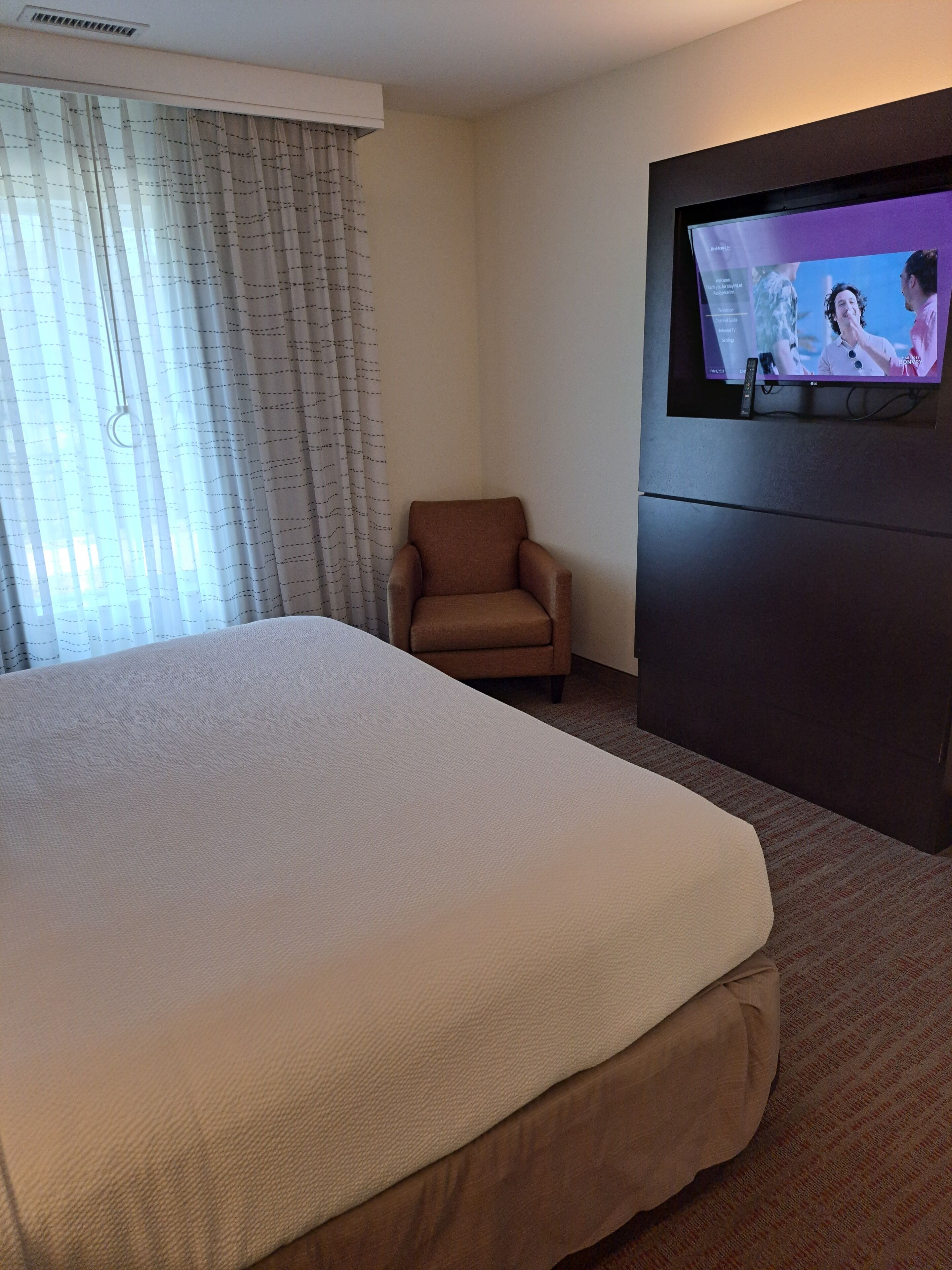 Una camera d'albergo con un letto grande, una poltrona marrone, una televisione a schermo piatto montata a parete e una finestra con tende trasparenti.