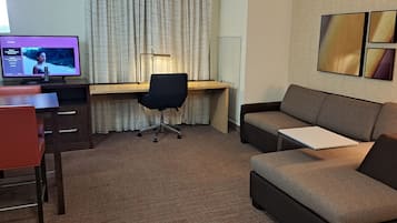 A hotel room with a desk, a black office chair, a brown sofa, a TV displaying a movie, and a wall with framed artwork.