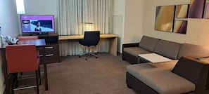A hotel room with a desk, a black office chair, a brown sofa, a TV displaying a movie, and a wall with framed artwork. - Residence Inn Columbia West/Lexington (West Columbia)