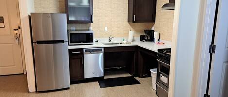 A compact kitchen with a stainless steel refrigerator, a microwave, a dishwasher, a sink, a stove, and dark wooden cabinets.