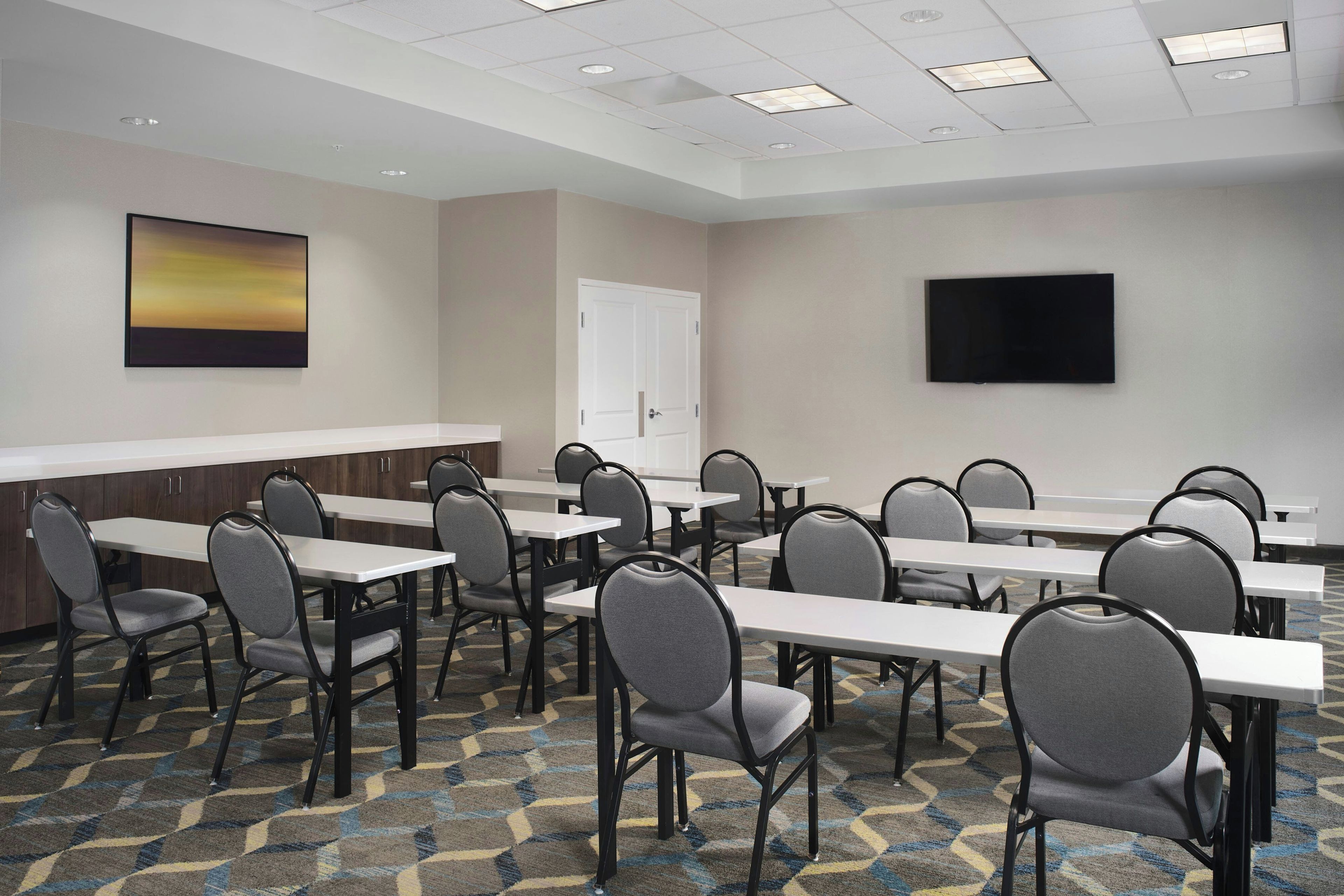 Une salle de conférence avec plusieurs tables et chaises, un téléviseur à écran plat accroché au mur et une porte au fond.