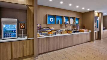 Un área de desayuno moderno en un hotel, con un mostrador de autoservicio, un refrigerador y diversas opciones de alimentos.