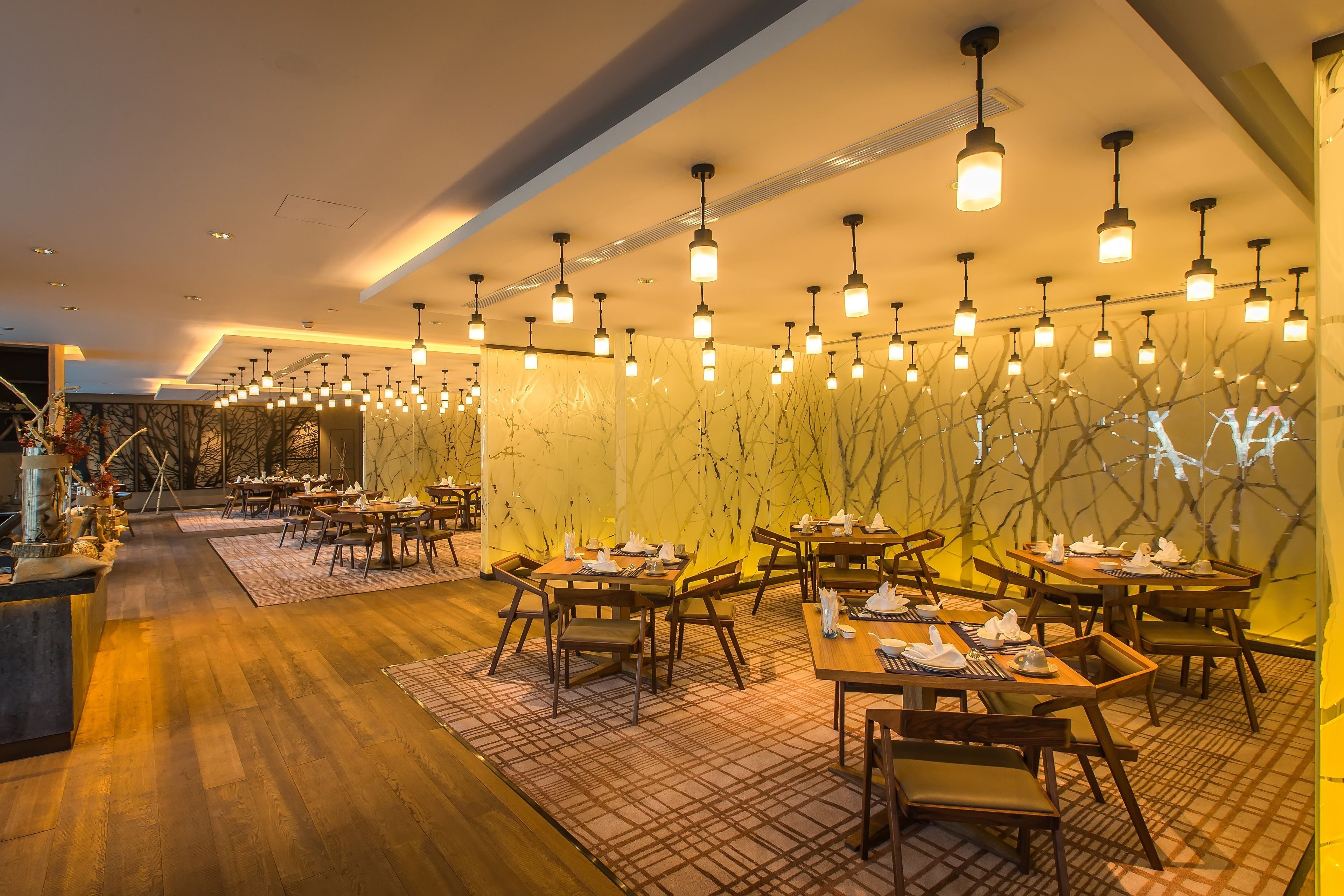 A restaurant with wooden tables and chairs, hanging pendant lights, and a marble wall with a tree design.