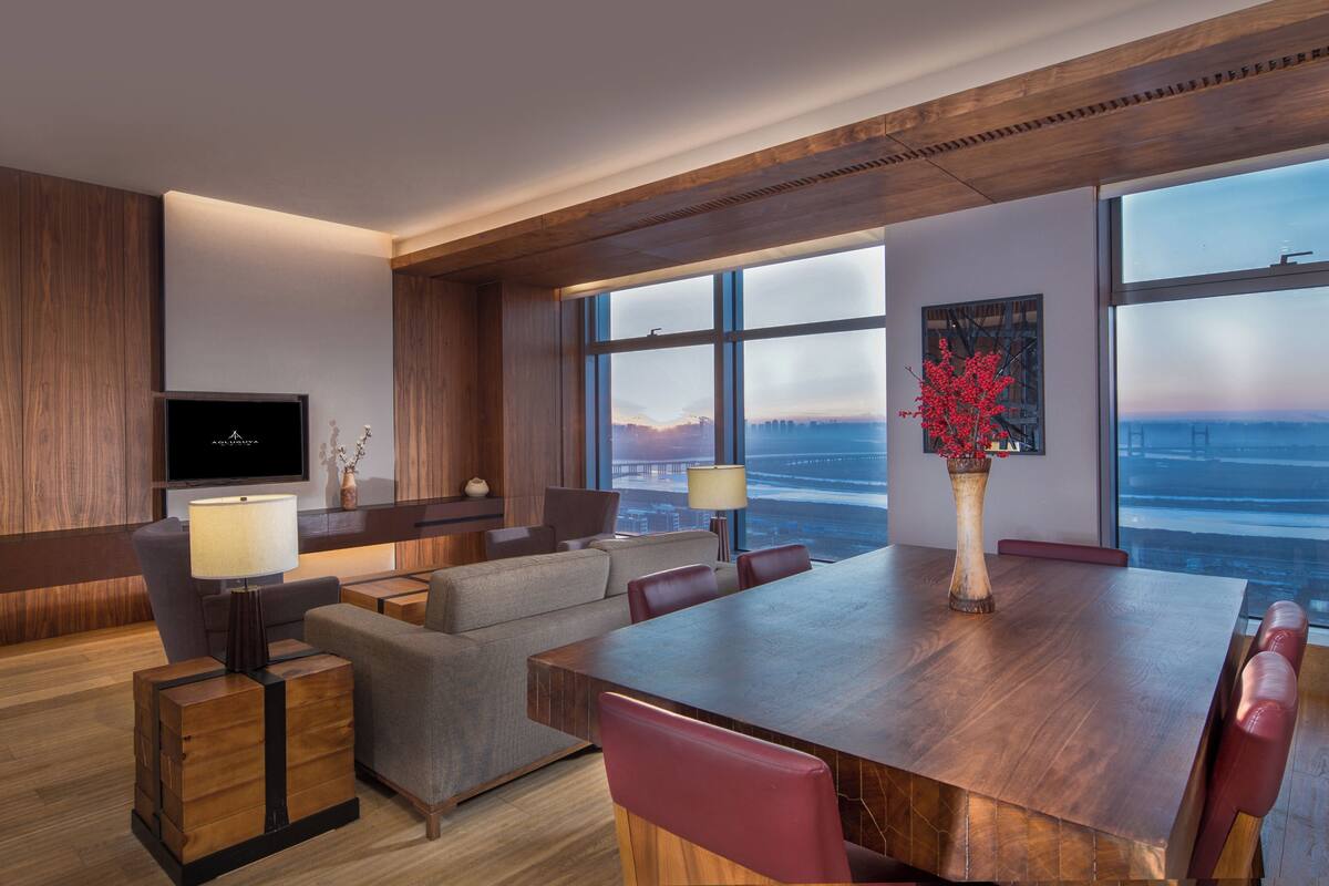Ein modernes Hotelzimmer mit großem Holztisch, bequemen Sitzgelegenheiten, einem Flachbildfernseher und einem großen Fenster mit Blick auf die Stadt.