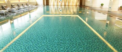 A large indoor swimming pool with tiled flooring and lounge chairs along the sides.