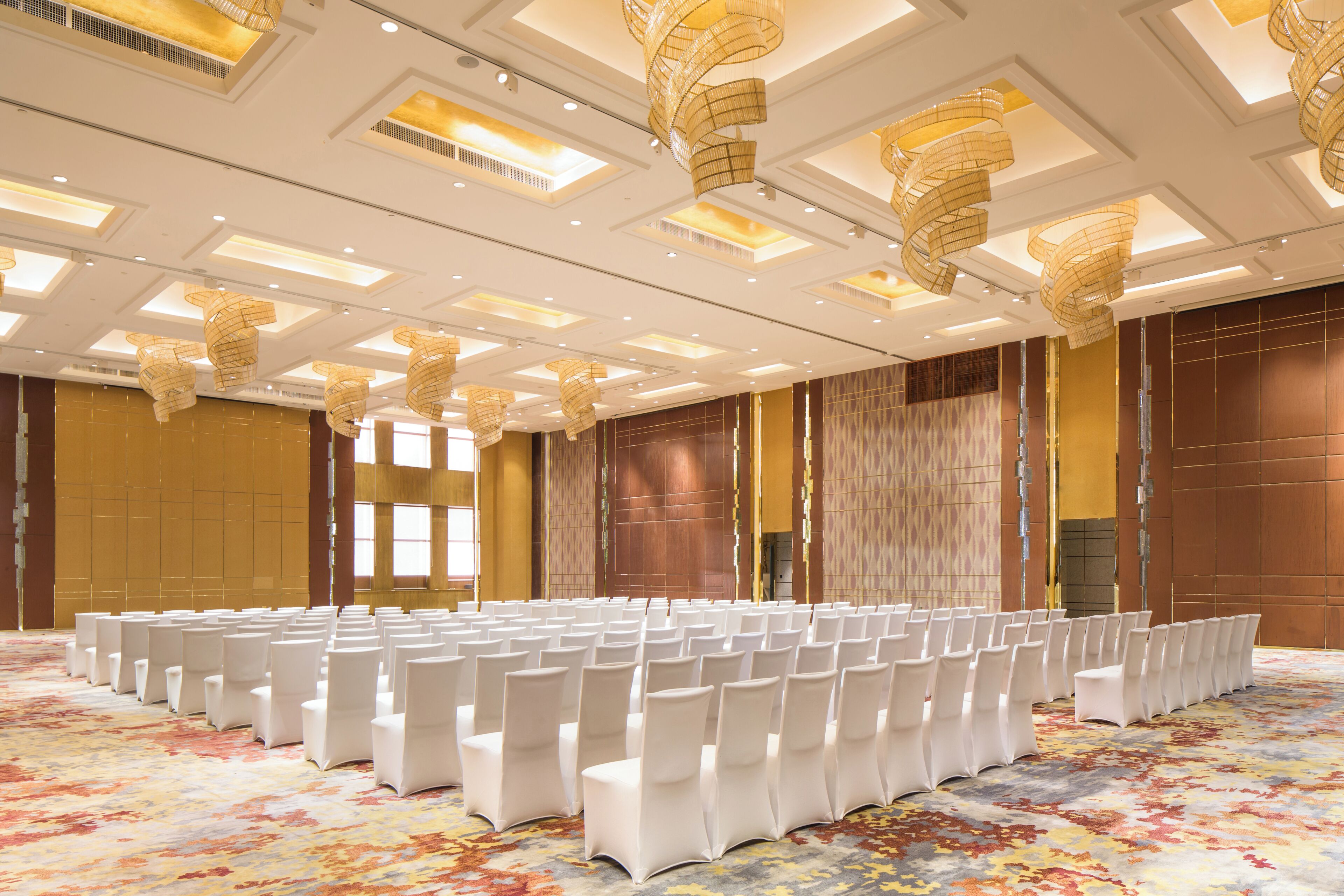 Une salle de conférence avec des rangées de chaises blanches, une moquette à motifs et un plafond orné de luminaires originaux.