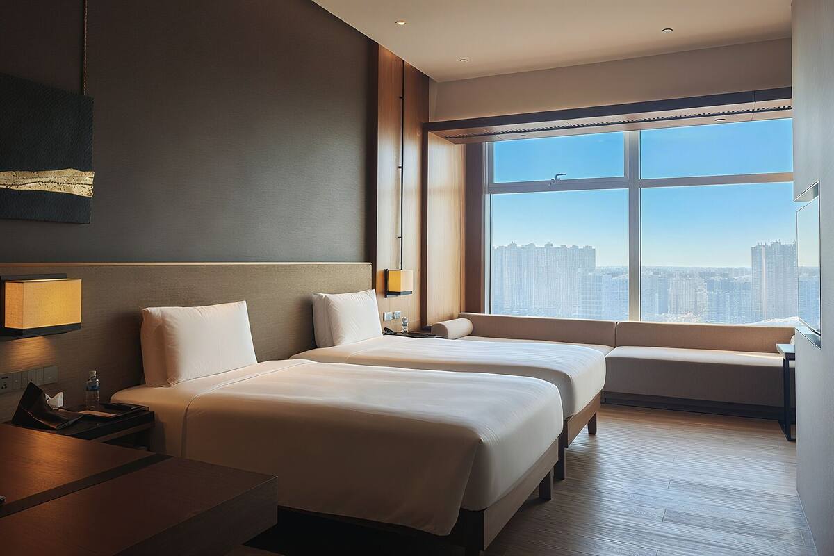 Ein Hotelzimmer mit zwei Betten, einem Schreibtisch, einem Sessel, einem großen Fenster mit Blick auf die Stadt und einer Wandlampe.