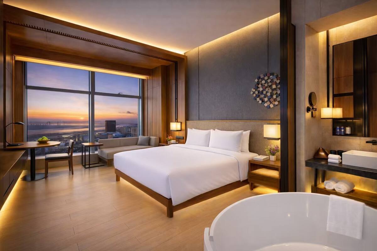 Ein modernes Hotelzimmer mit einem großen Bett, einer Sitzecke, Blick auf die Stadt bei Sonnenuntergang, einem Badezimmer mit Badewanne und einem Schreibtisch mit Lampe.