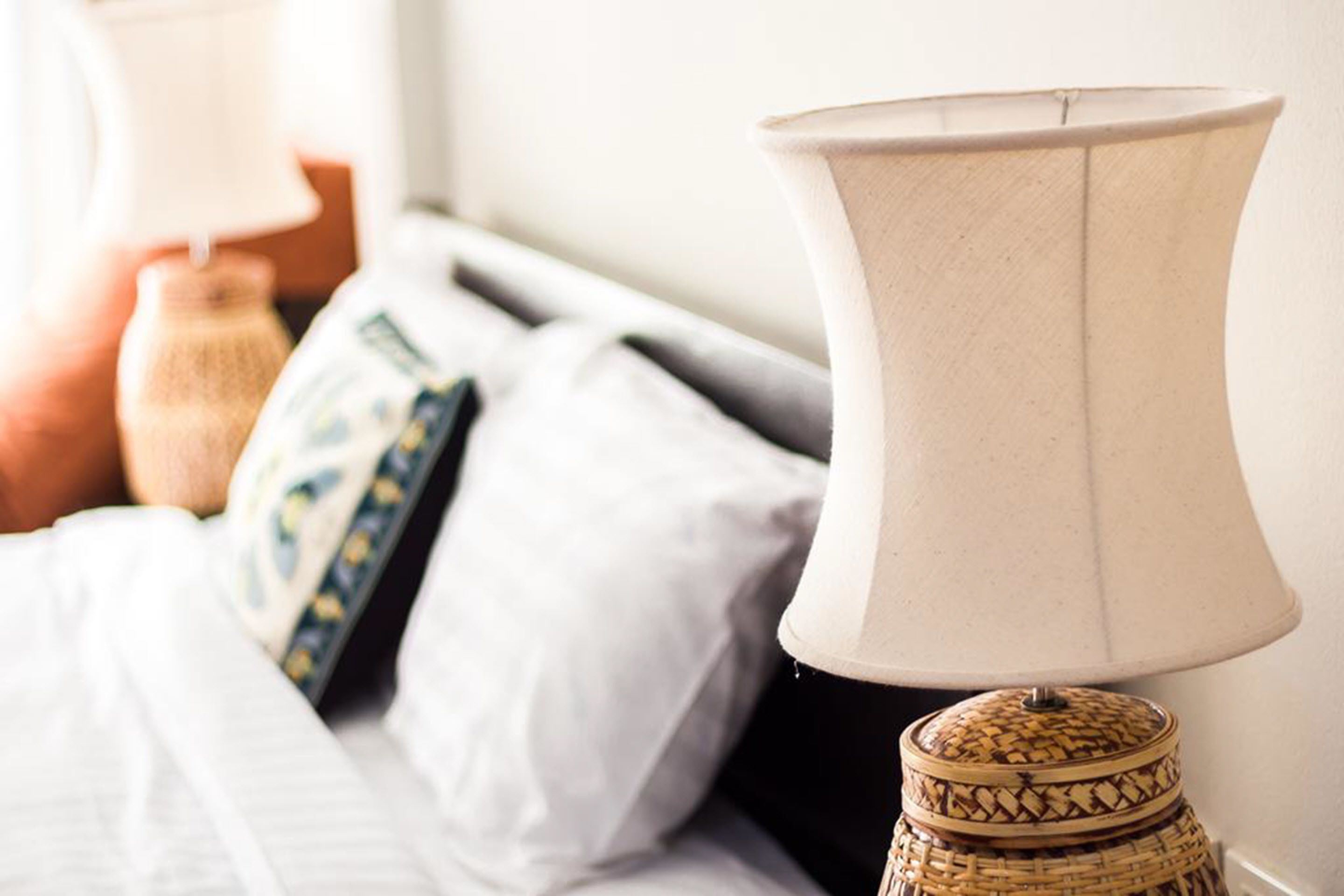A bed with a white comforter, a decorative pillow, and a lamp with a woven base.